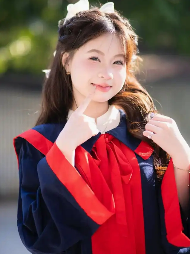 Cô gái trẻ mặc áo cử nhân màu xanh đỏ mỉm cười đặt ngón tay lên môi duyên dáng